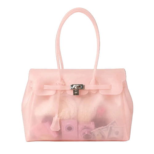 A translucent light pink handbag with a structured rectangular shape, dual top handles, a front flap with a tie detail, and a silver clasp, shown on a clean white background. Inside, pink and beige accessories are partially visible through the transparent material.